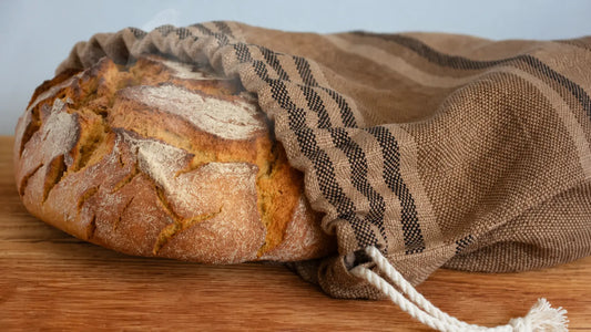Pane rustico appena sfornato, parzialmente avvolto in un sacchetto di stoffa marrone su un tavolo in legno, con un leggero fumo che sale dalla crosta dorata.