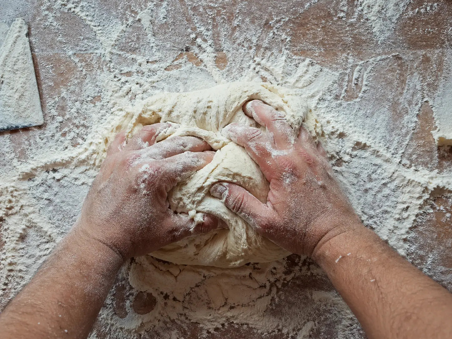 Mani di un panificatore che impastano il pane su un banco da lavoro in legno ricoperto di farina.