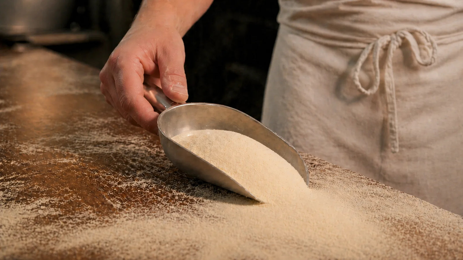 Panificatore che usa un dosatore in metallo che raccoglie farina di semola su un banco di legno in forno artigianale.