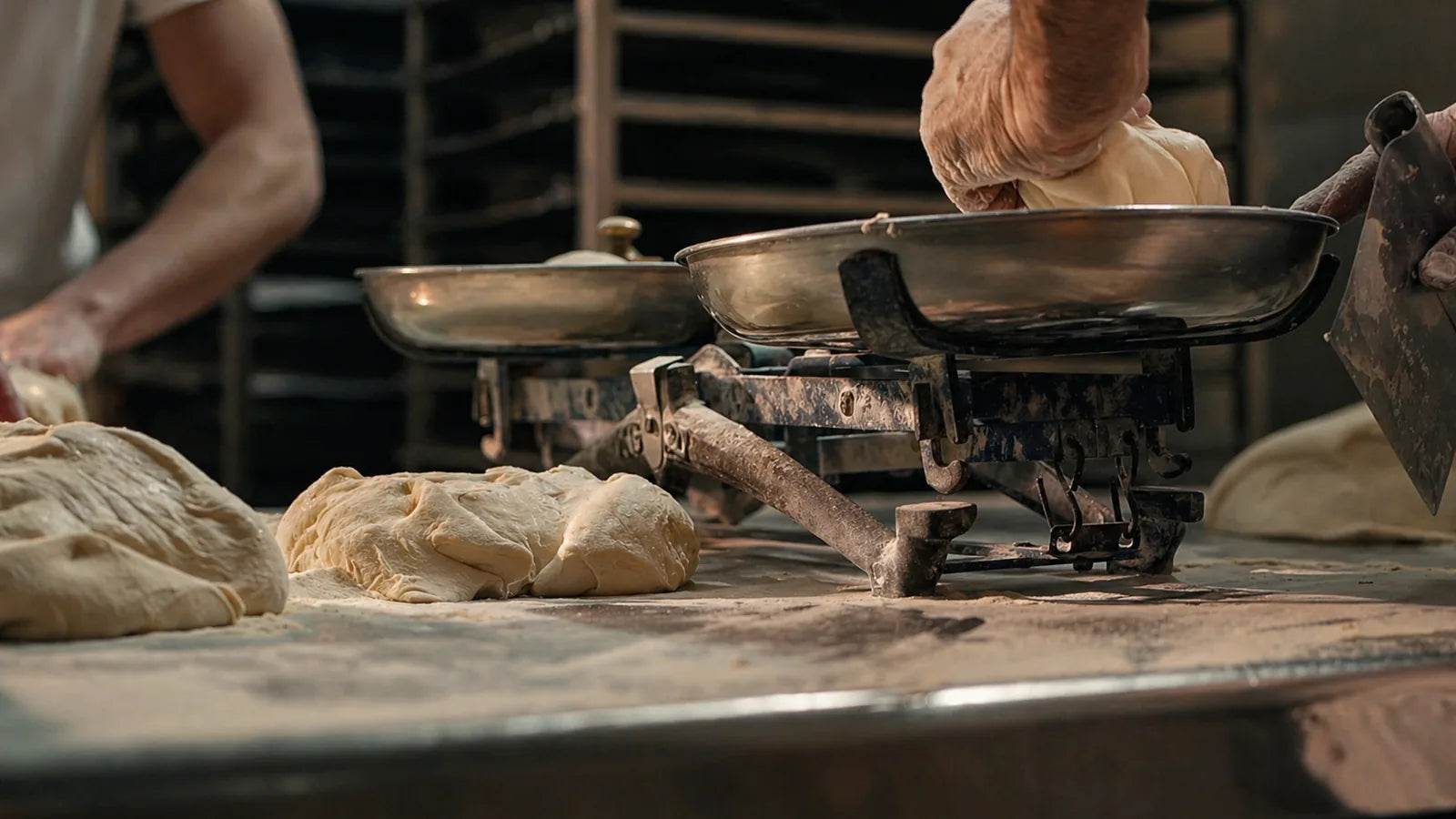Impasti di pane su un banco infarinato, con bilancia da panificio e mani dei due fornai al lavoro in laboratorio.