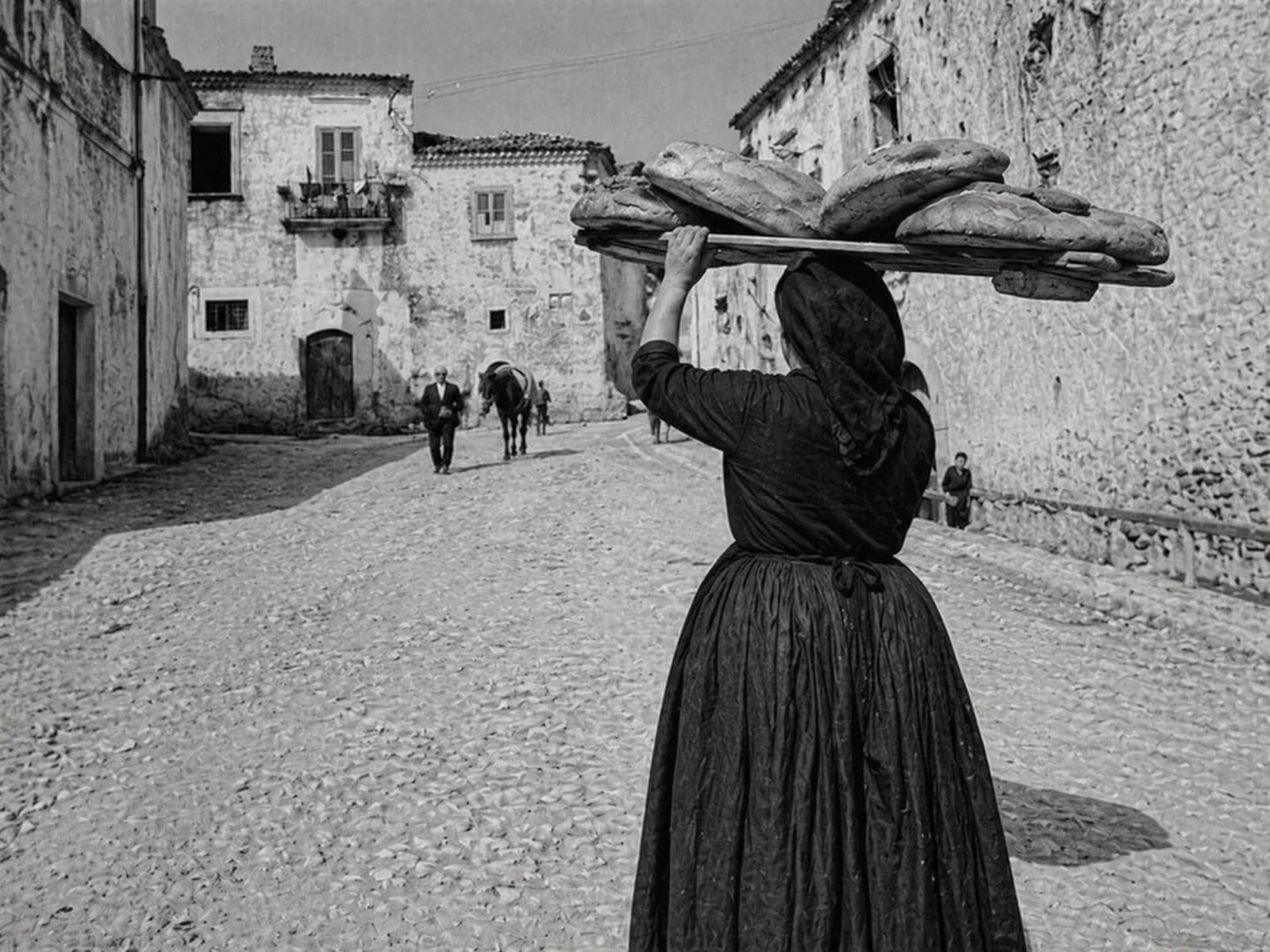 Donna che trasporta il pane su una tavola nei Sassi di Matera, in una scena storica in bianco e nero.