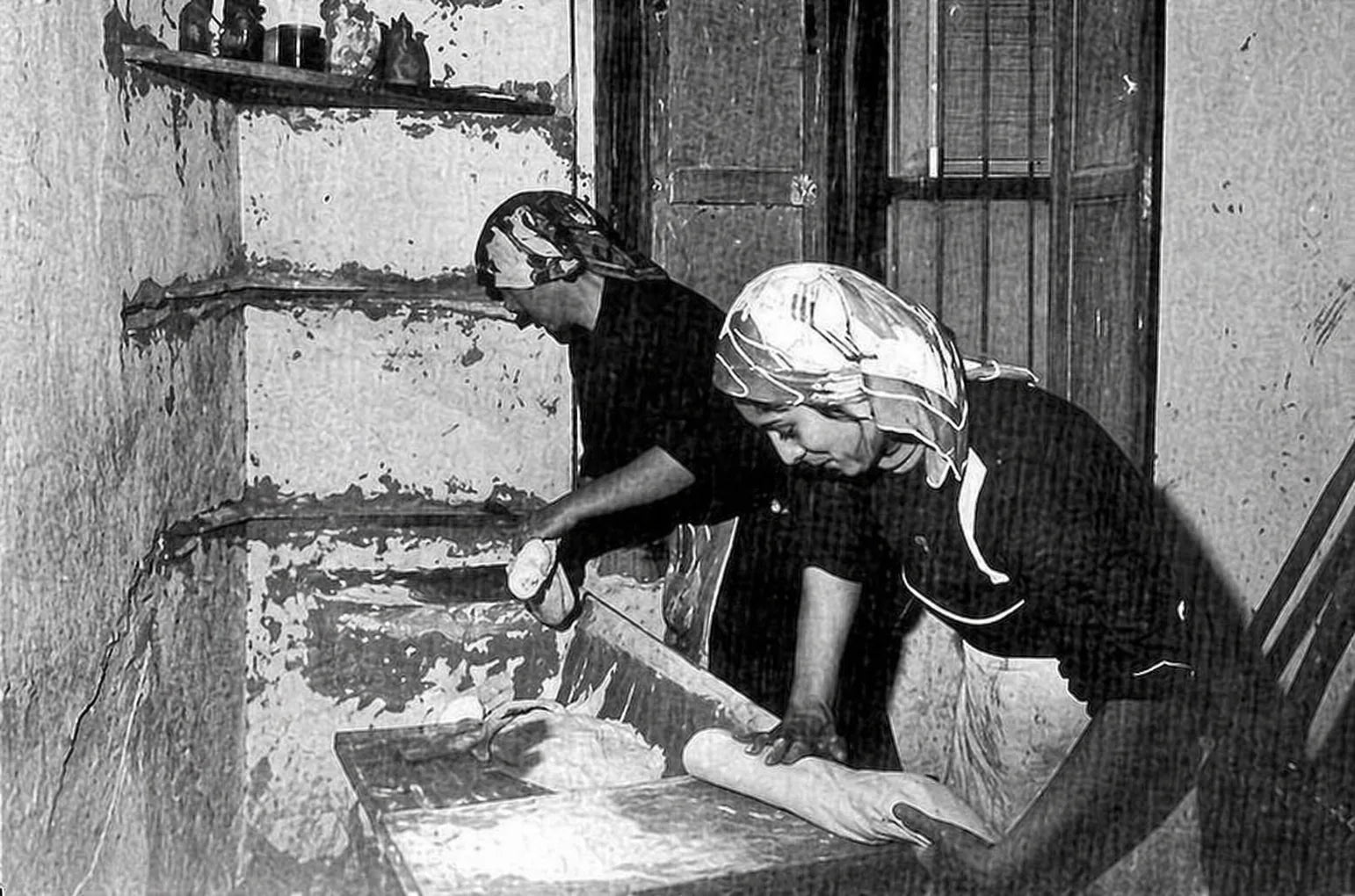 Due donne impastano e lavorano il pane in un ambiente domestico tradizionale dei Sassi di Matera, in una scena storica in bianco e nero.